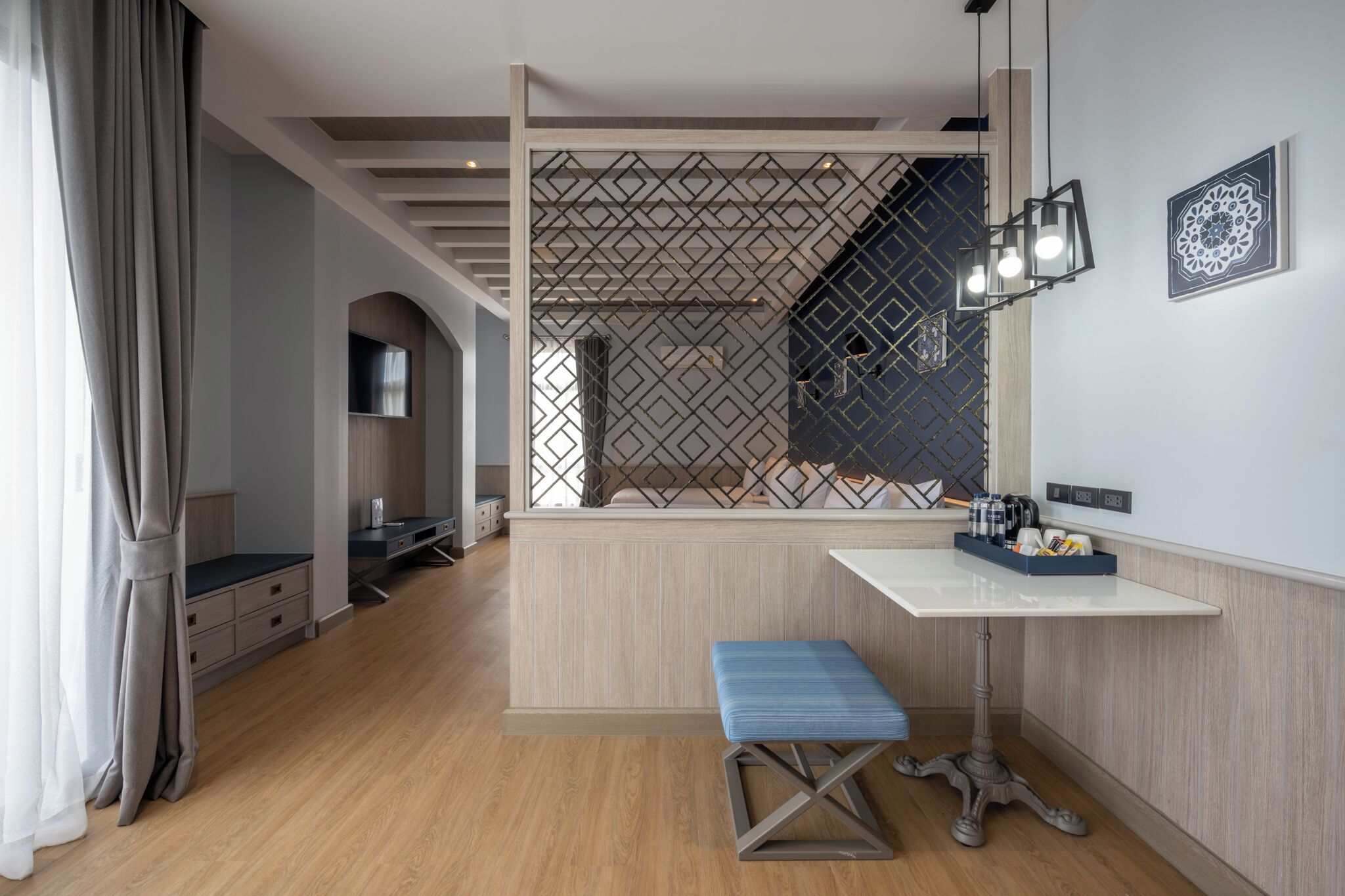 Una habitación de hotel moderna con piso de madera, un escritorio blanco con una bandeja azul, un banco azul y un panel decorativo en la pared.