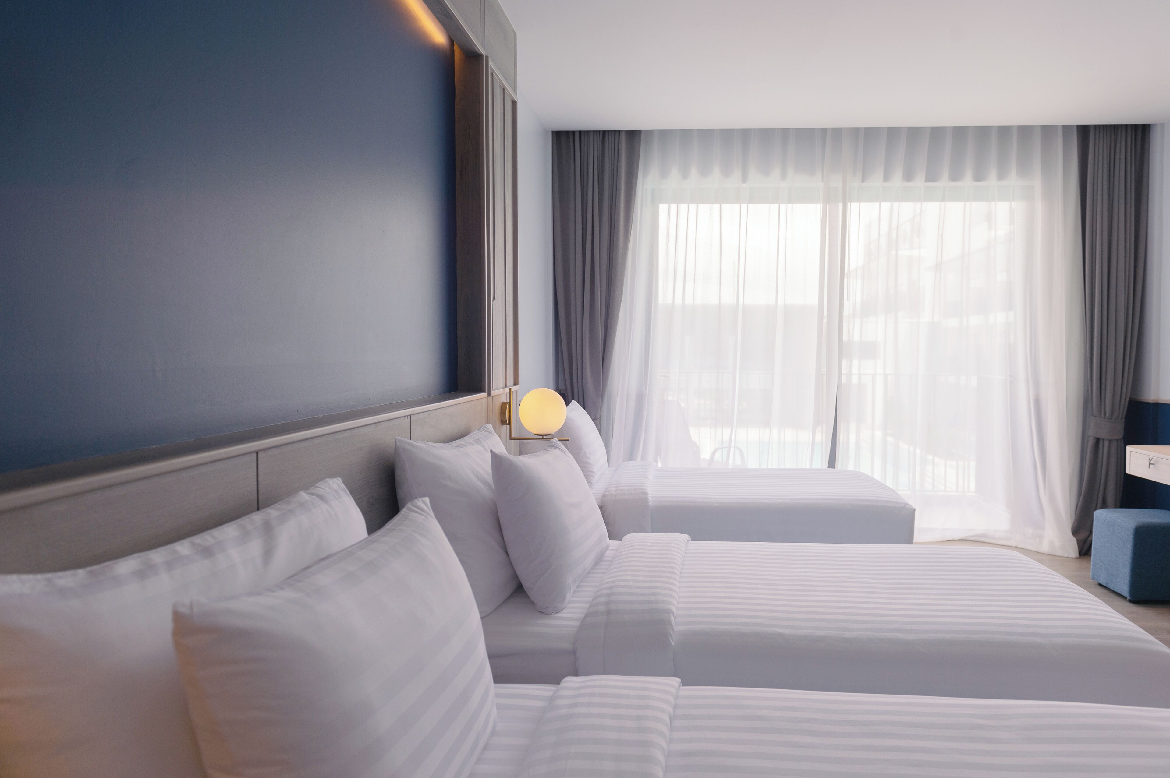 A hotel room with two beds, a bedside table with a lamp, and a large window with sheer curtains.