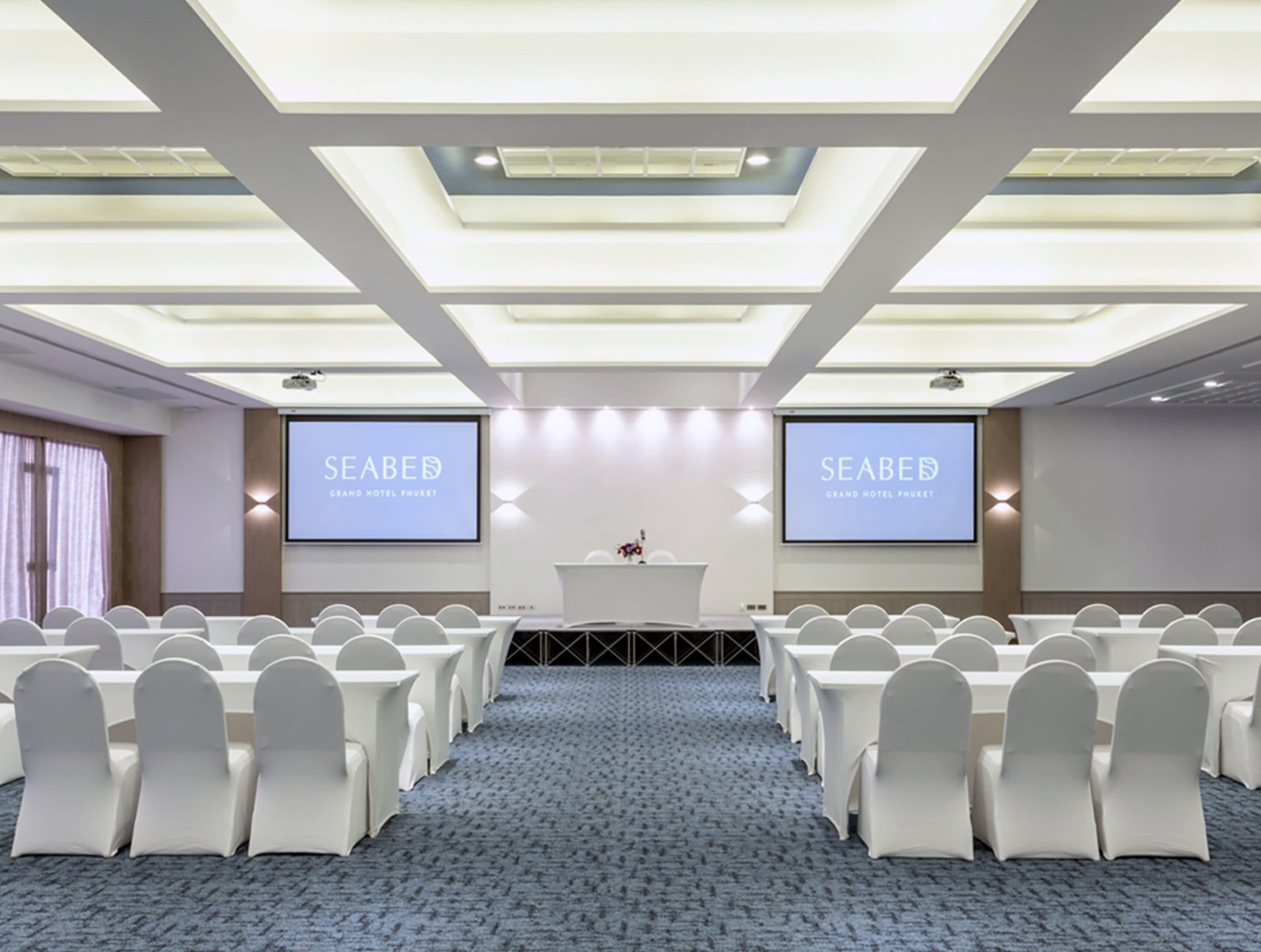 A conference room with a stage, two screens displaying 'SEABED GRAND HOTEL PHUKET', and rows of white chairs.