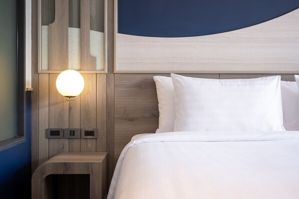 Una cama bien hecha con sábanas blancas, una mesita de noche con lámpara y una lámpara de pared.