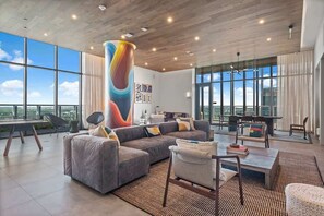 A spacious living area with a large sectional sofa, a coffee table, and a dining table. There are multiple chairs, a tall pillar with a colorful design, and large windows offering a view of the cityscape.