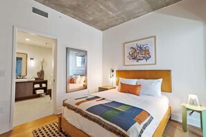 A bedroom with a bed, a nightstand, a lamp, a mirror, and a bathroom visible through an open door.
