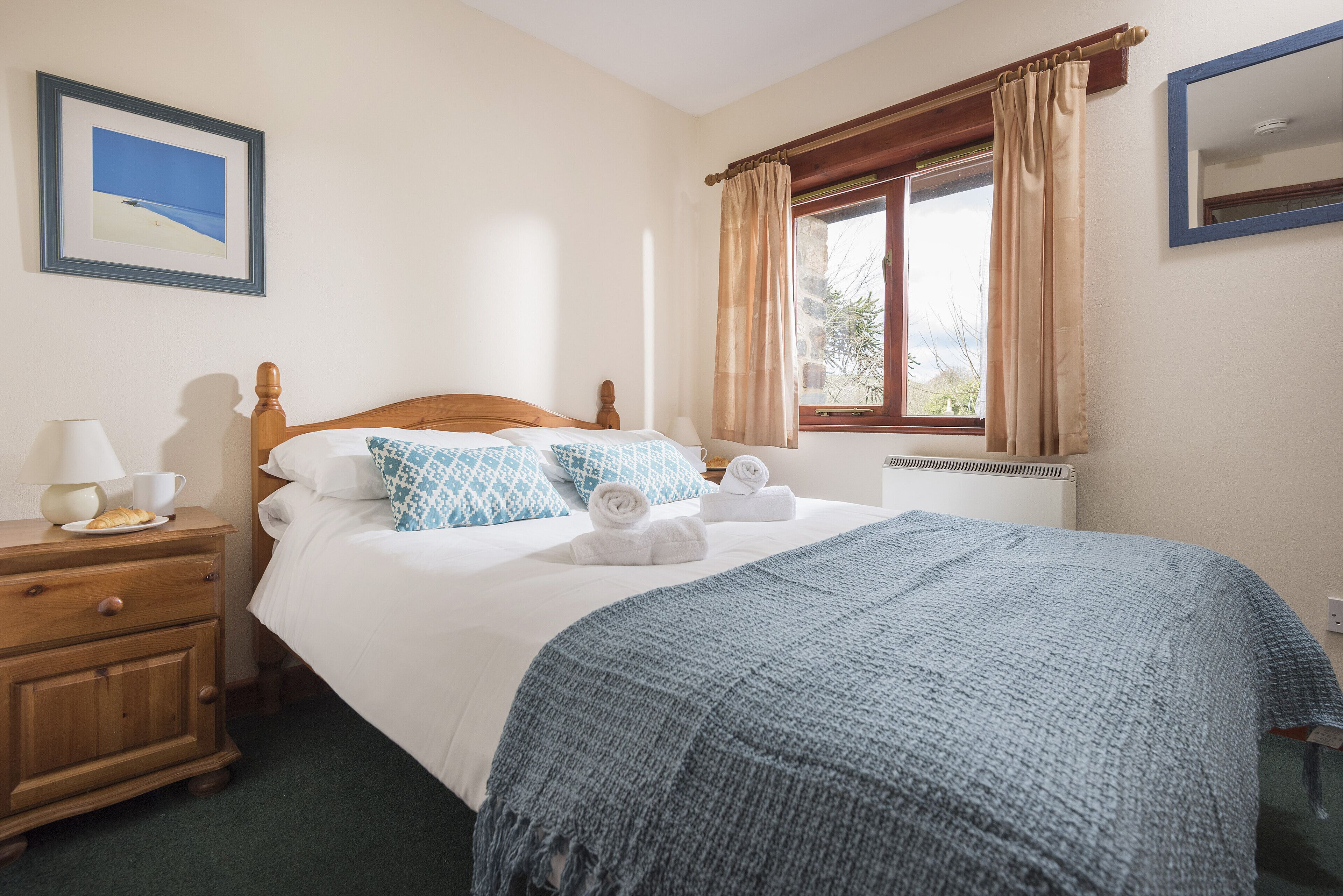 A neatly made bed with a blue blanket, a wooden nightstand with a lamp, and a framed picture on the wall.