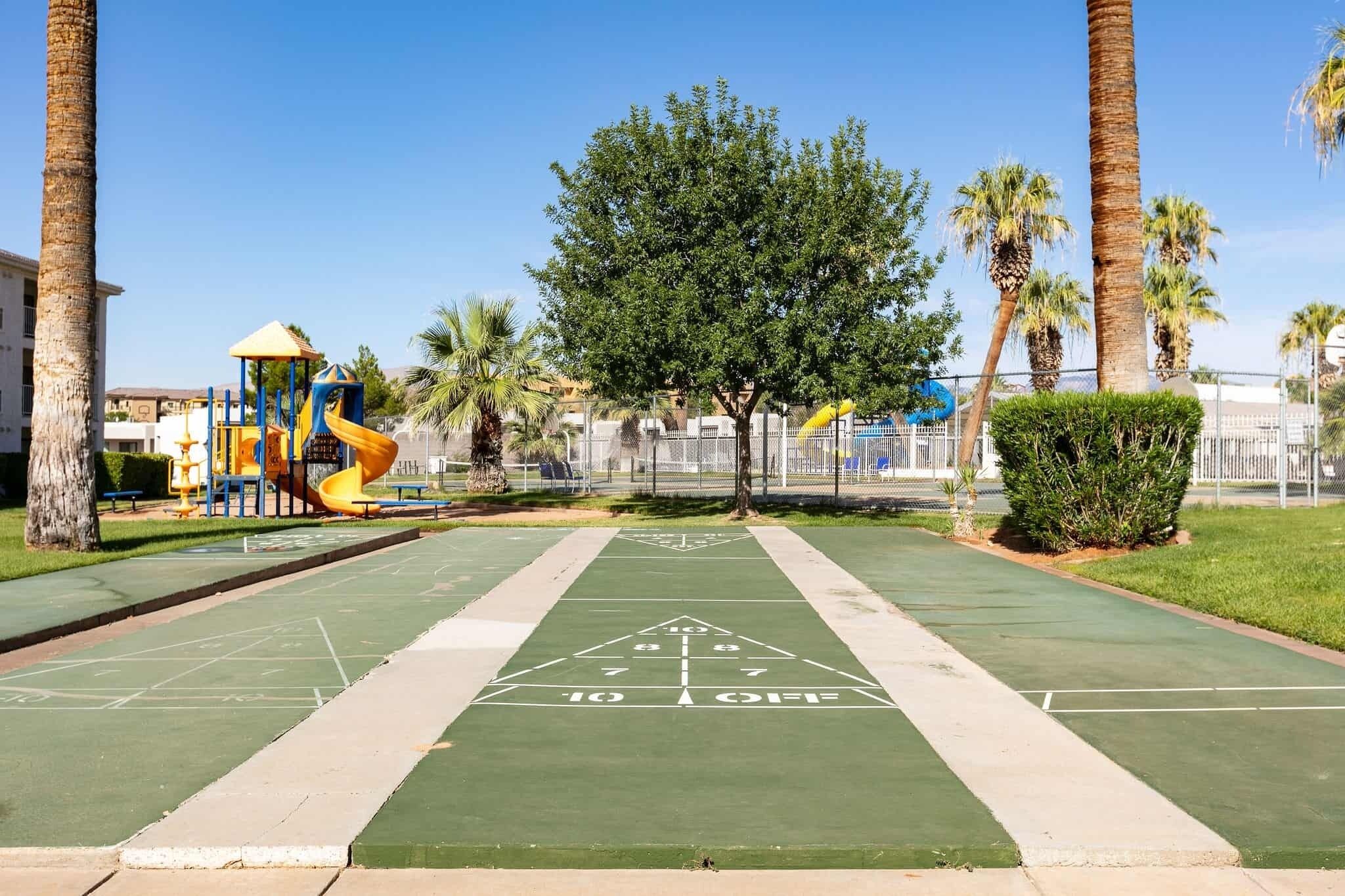 A park with a playground, a green shuffleboard court, and palm trees.
