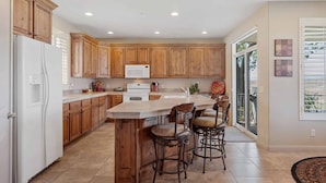 A kitchen with wooden cabinets, a white refrigerator, a microwave, and a large island with bar stools. - Tuscan Desert Vista #2001 by RedAwning (St. George)