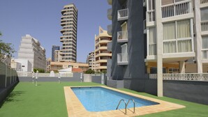 A swimming pool with a green lawn area, surrounded by multi-story residential buildings. - A77 HERNANDO I 3º - G (Calpe)