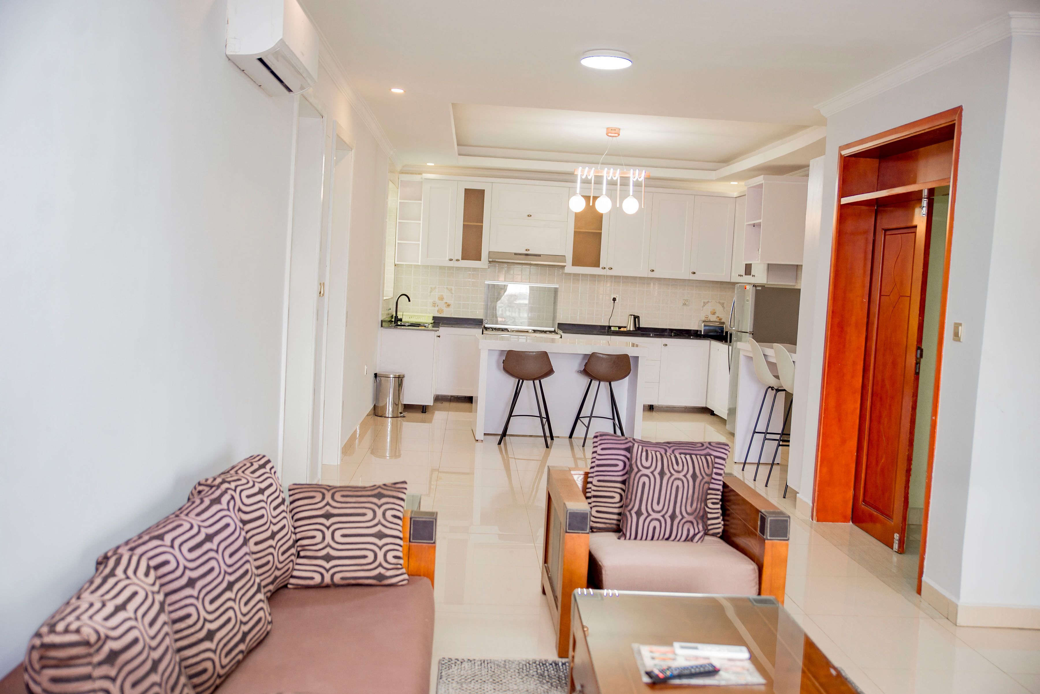 Un salon moderne avec un canapé et un fauteuil, une table basse, et une cuisine en arrière-plan avec des meubles blancs et un coin repas.