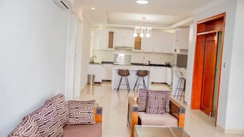 Un salon moderne avec un canapé et un fauteuil, une table basse, et une cuisine en arrière-plan avec des meubles blancs et un coin repas.