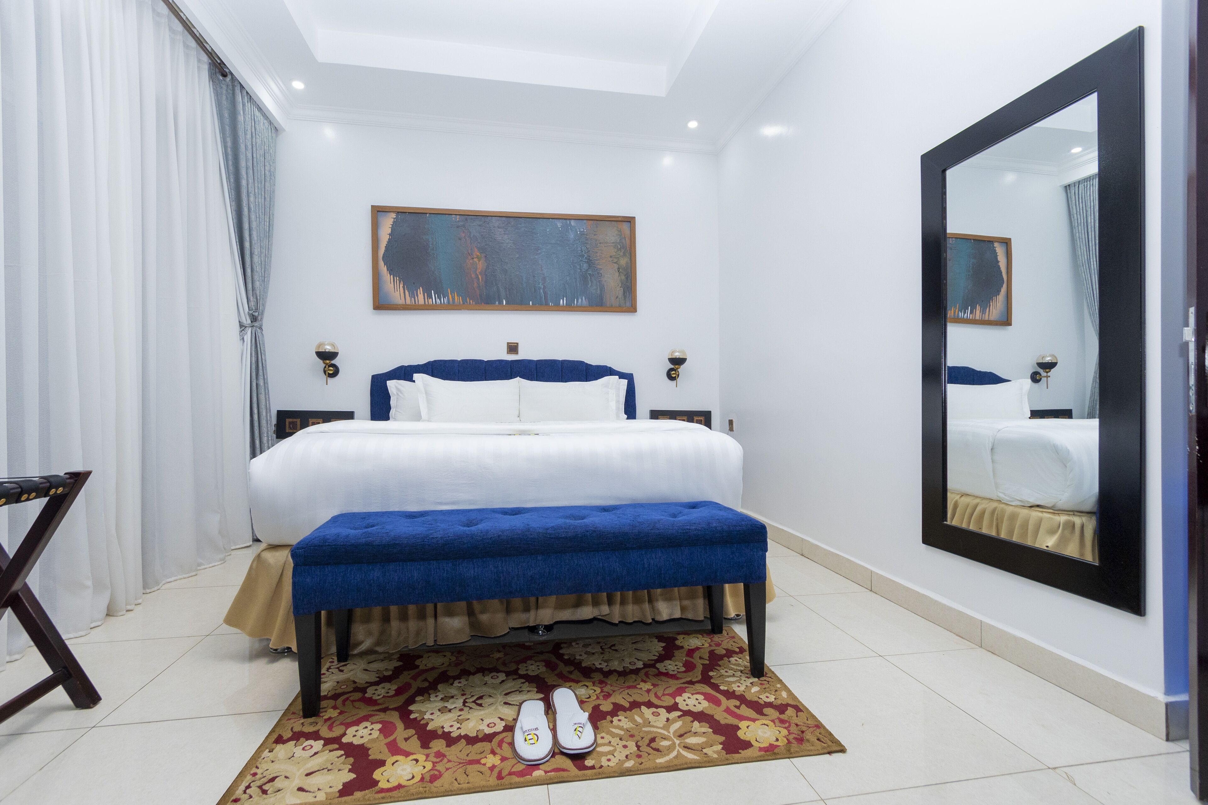 A bedroom with a bed, a bench, a pair of slippers, a rug, and a large mirror.