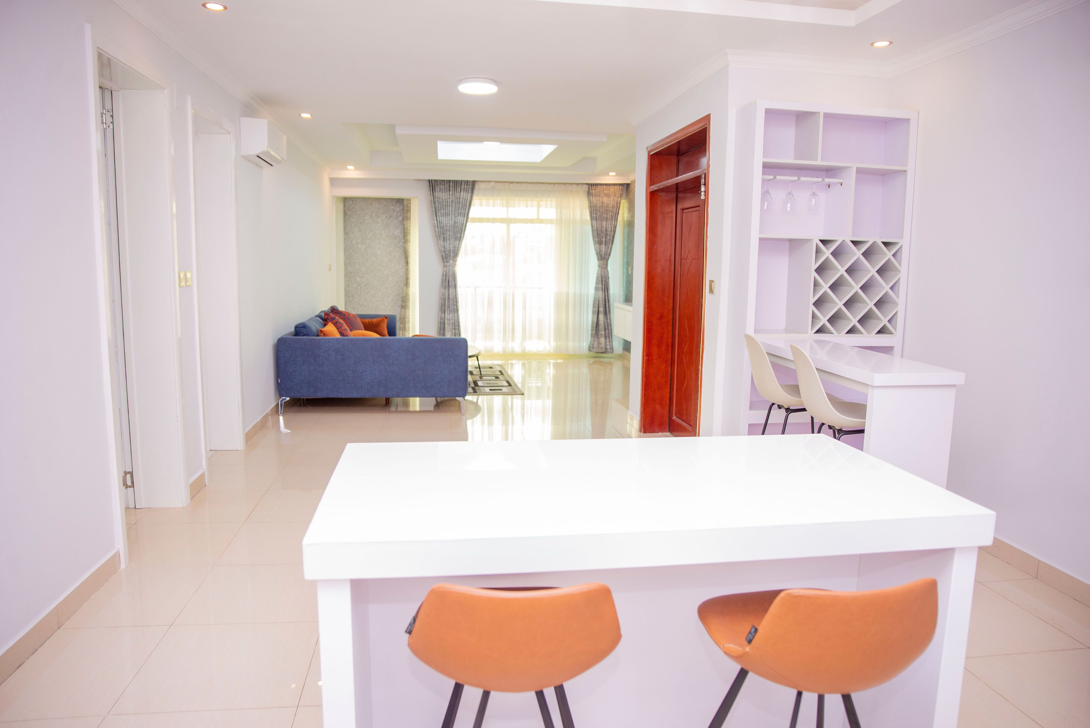 Un salon moderne avec un canapé bleu, une table à manger blanche et deux chaises orange, ainsi qu’une porte en bois.