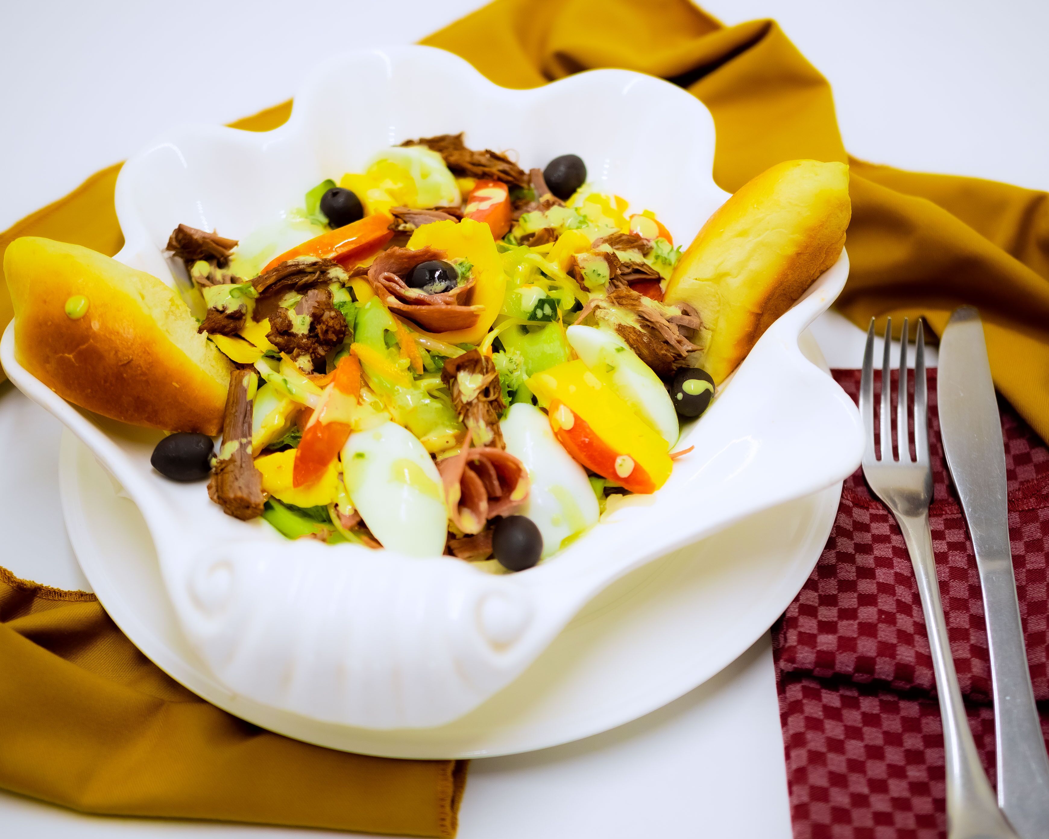 A white plate with a salad, two bread rolls, and a fork and knife on a red and yellow napkin.
