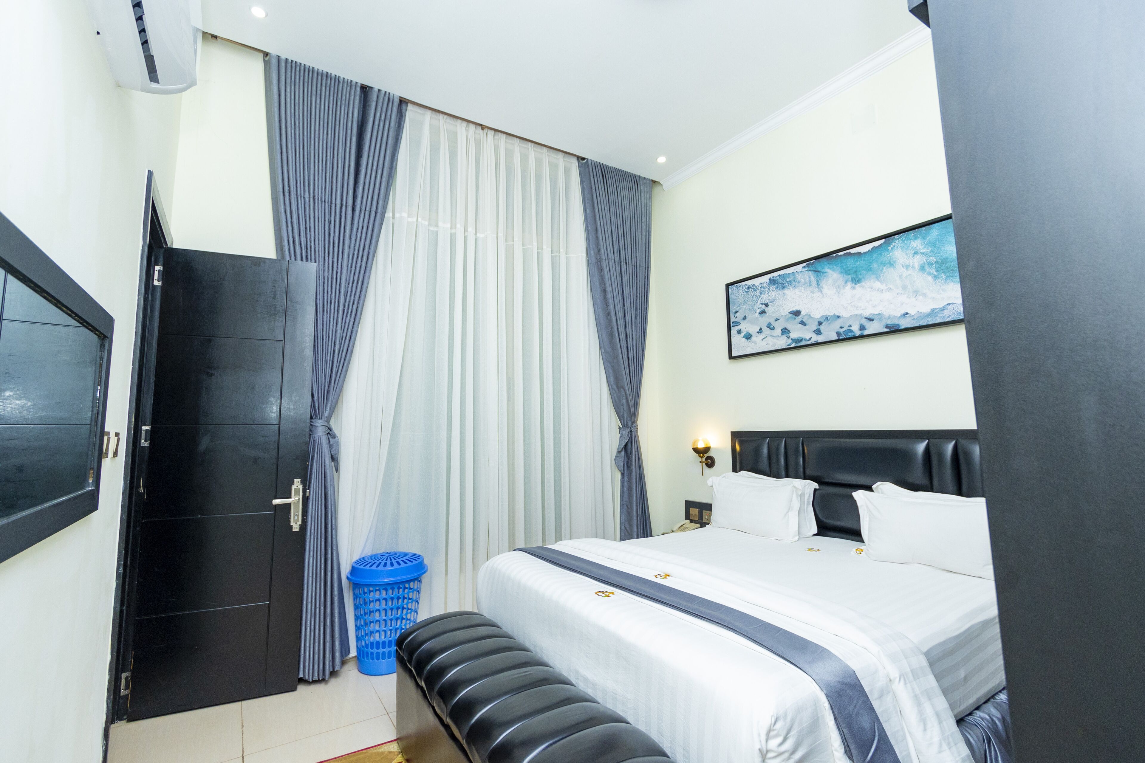 A hotel room with a bed, a bench, a trash bin, a door, and a painting on the wall.