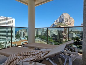 Un balcón con una silla de descanso y una toalla a rayas, con vistas a un paisaje urbano y una colina rocosa destacada.