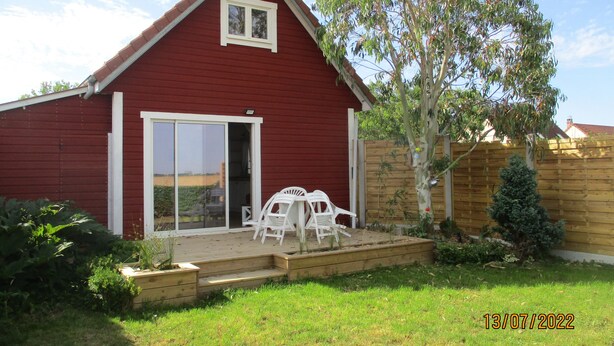 Un bâtiment rouge avec une fenêtre blanche et une porte-fenêtre coulissante, une terrasse en bois avec des chaises blanches et un petit arbre.