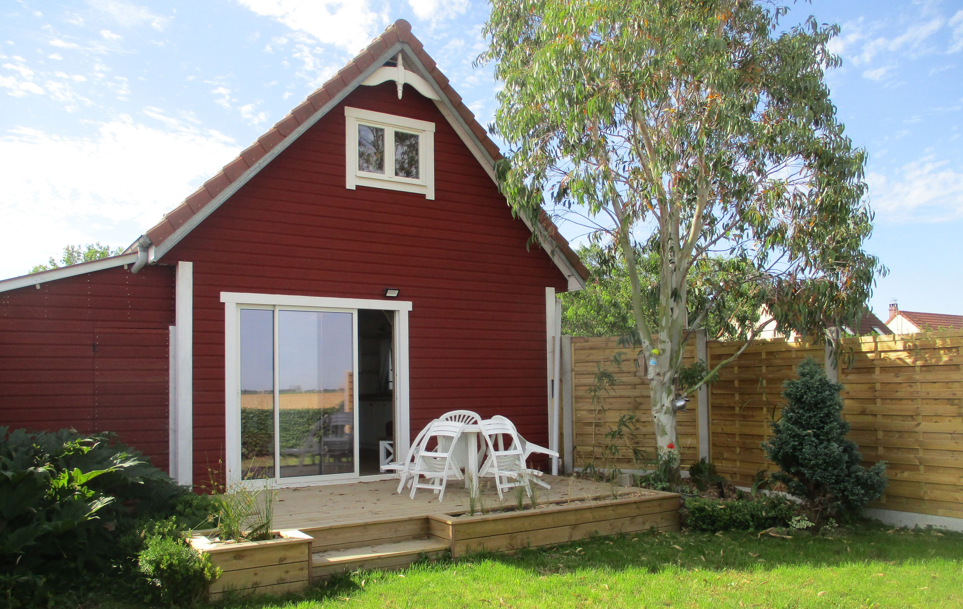 A red building with a white window and sliding glass door, a wooden deck with white chairs, and a small tree.