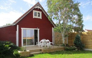Un bâtiment rouge avec une fenêtre blanche et une porte-fenêtre coulissante, une terrasse en bois avec des chaises blanches et un petit arbre.