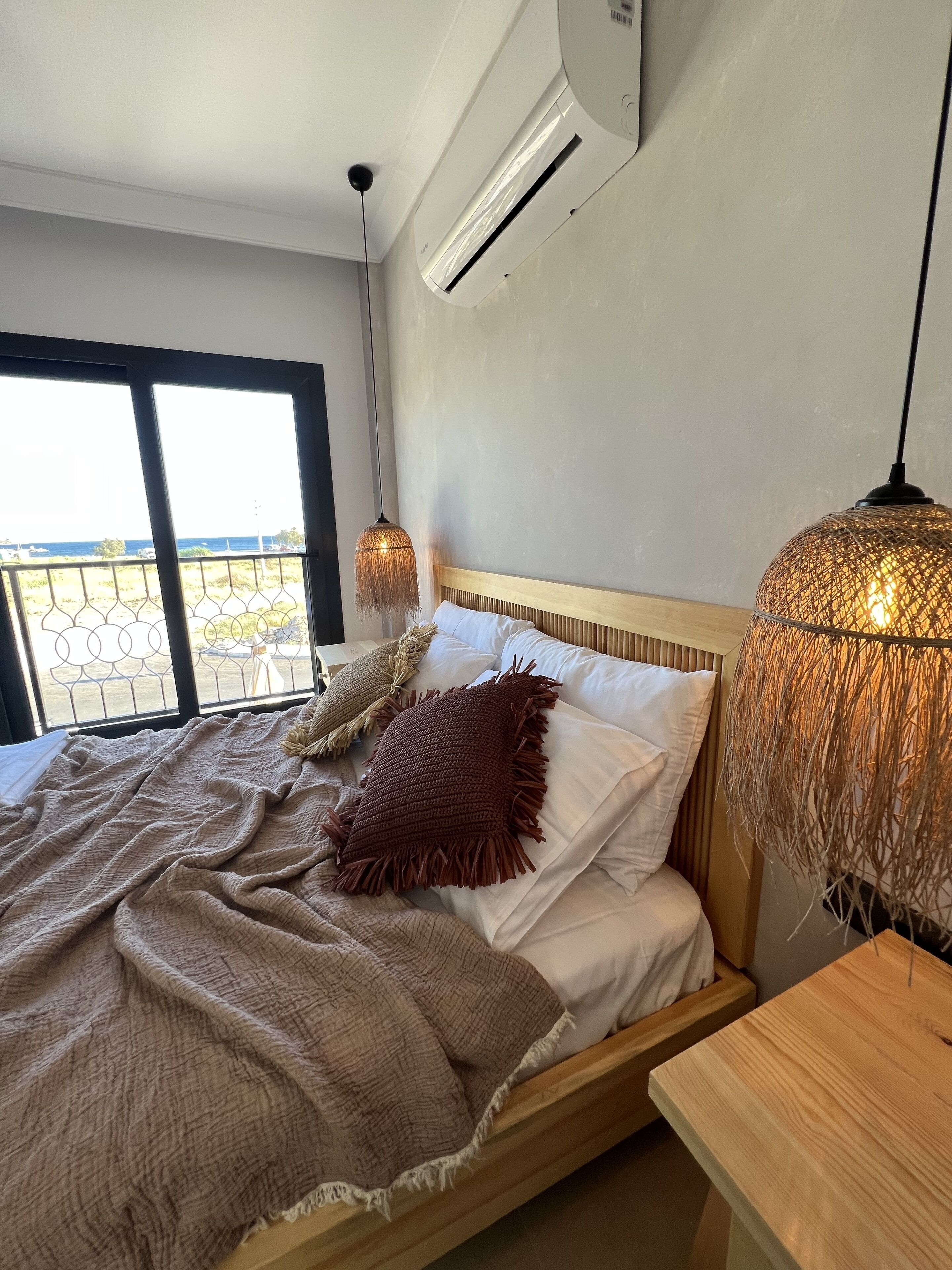 A bedroom with a bed, a wooden headboard, a large window with a view, and a hanging light fixture.