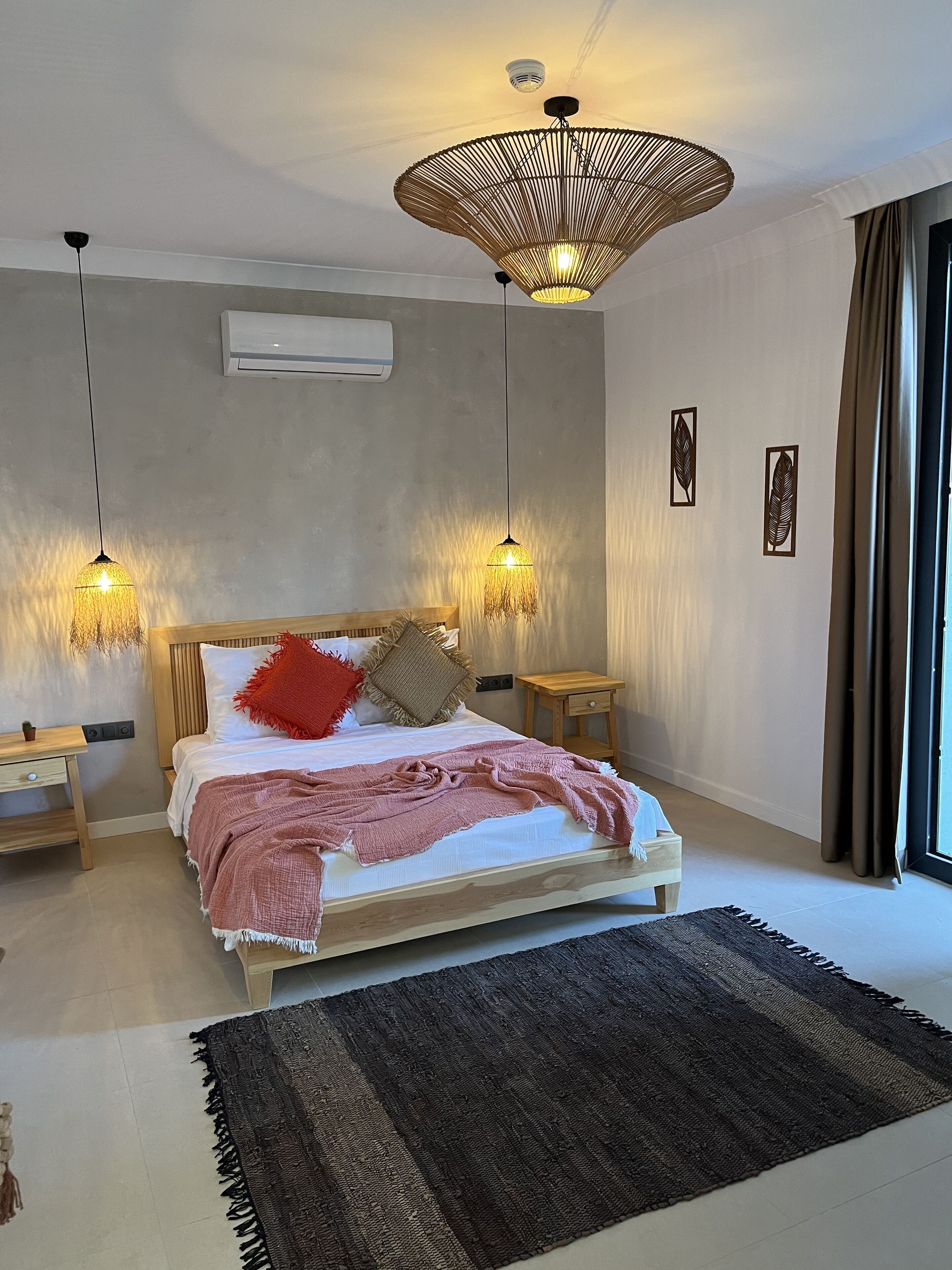 A modern bedroom with a large bed, two bedside tables, a wooden headboard, a large woven light fixture, and a view of the outdoors through a window.