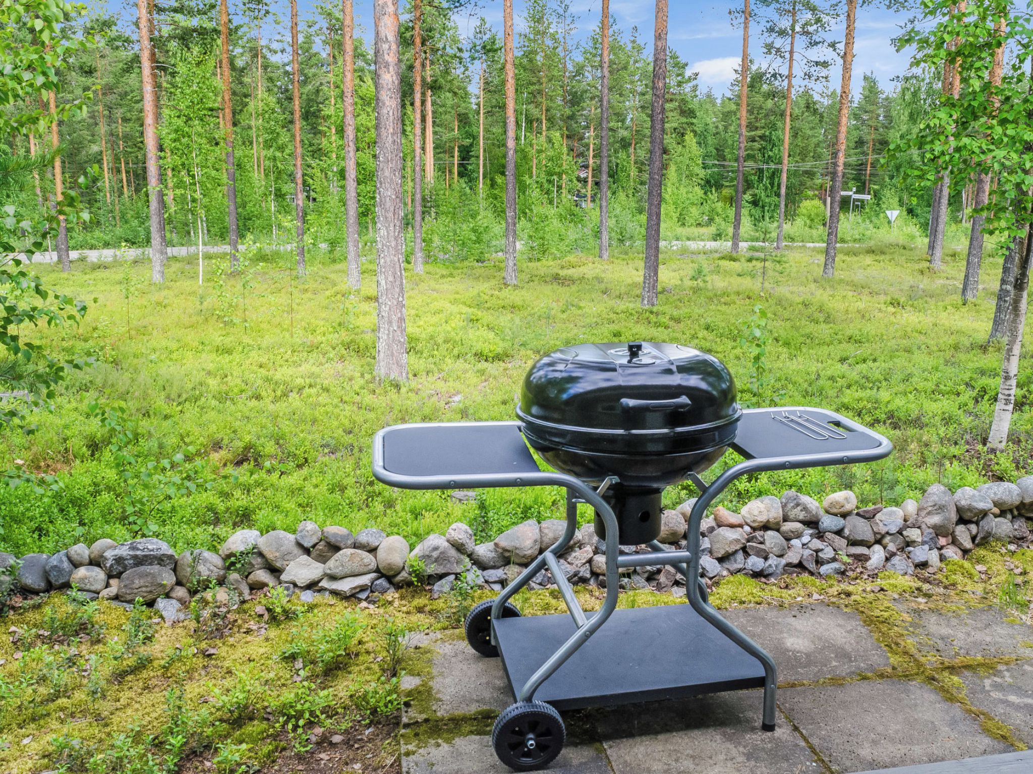 An outdoor grill with a cart, set on a stone path with a grassy area and trees in the background.