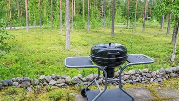 An outdoor grill with a cart, set on a stone path with a grassy area and trees in the background.