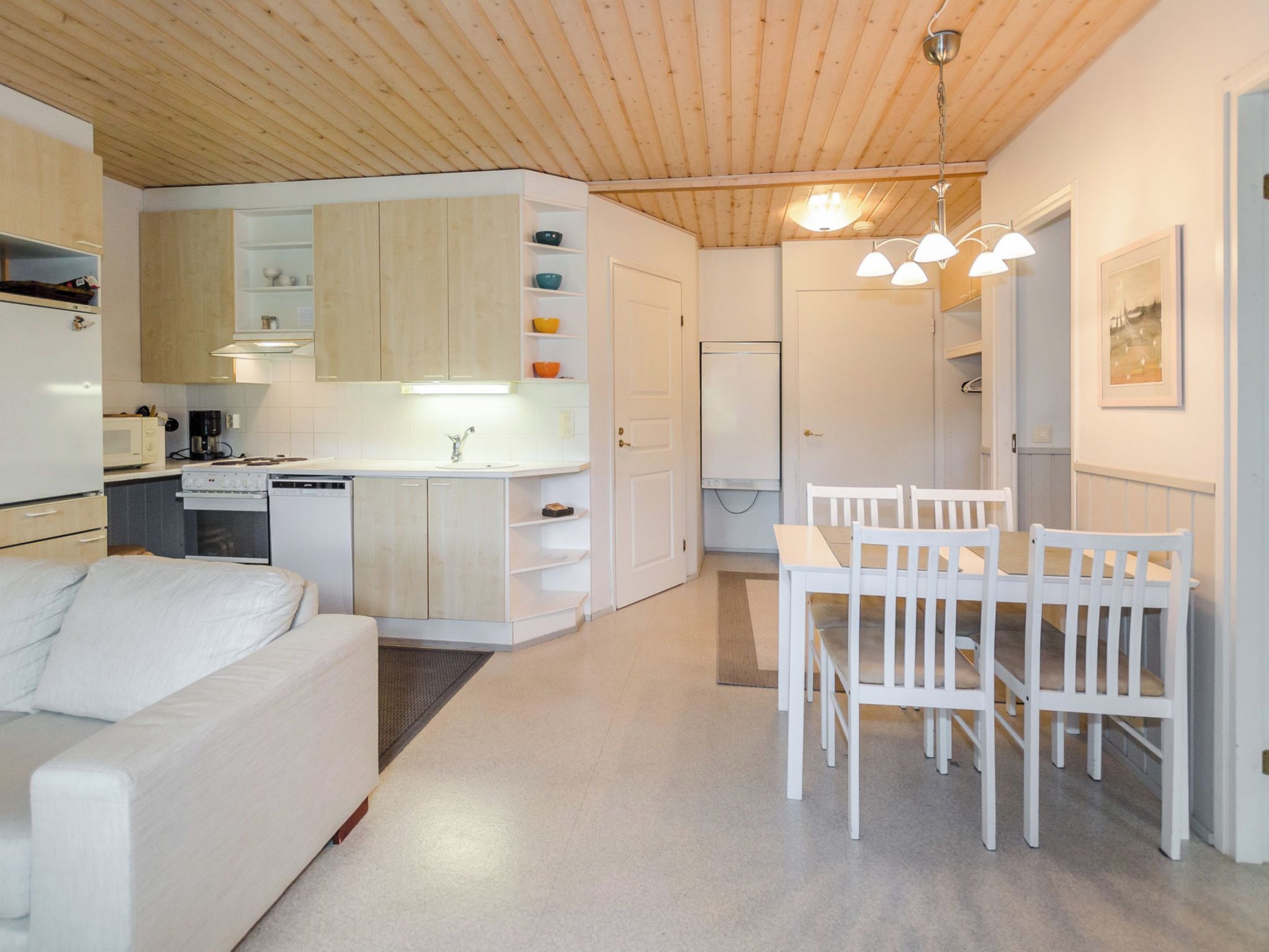 A modern kitchen with white cabinets, a dining table with chairs, and a white sofa in a bright room with wooden ceiling beams.