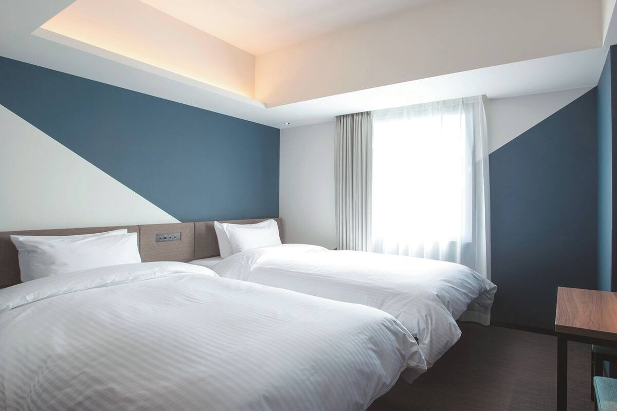 A hotel room with two beds, a wooden headboard, a small table, and a window with curtains.