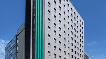 Um hotel moderno com uma faixa verde marcante, vários andares e céu azul ao fundo.