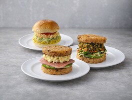 Three sandwiches on white plates against a grey background.