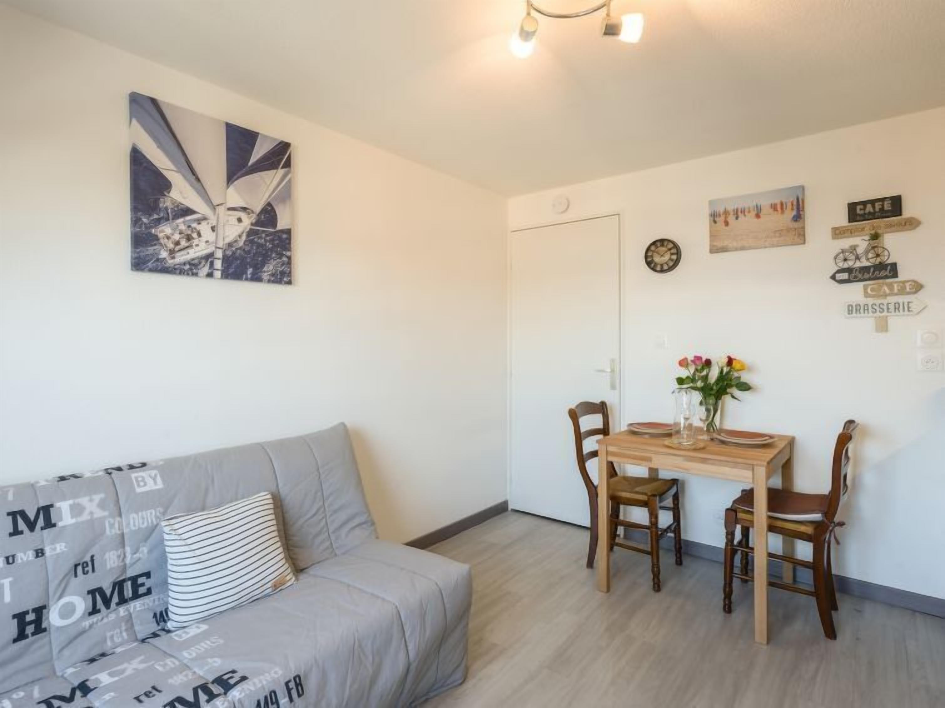 A cozy living room with a grey sofa, a wooden dining table with chairs, and a wall with various signs and a clock.