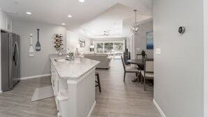 A modern kitchen with a central island, a dining area with a table and chairs, and a living room with a sofa and a large window.