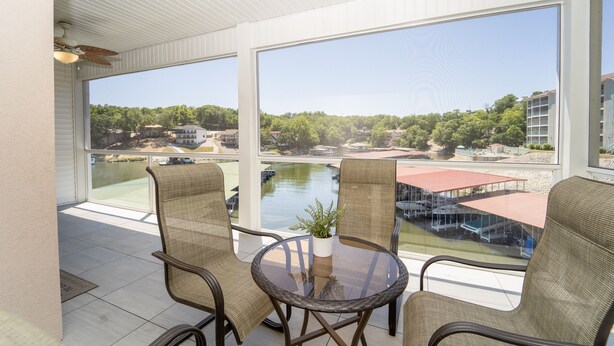 A balcony with wicker chairs, a glass table, and a view of a lake and buildings.