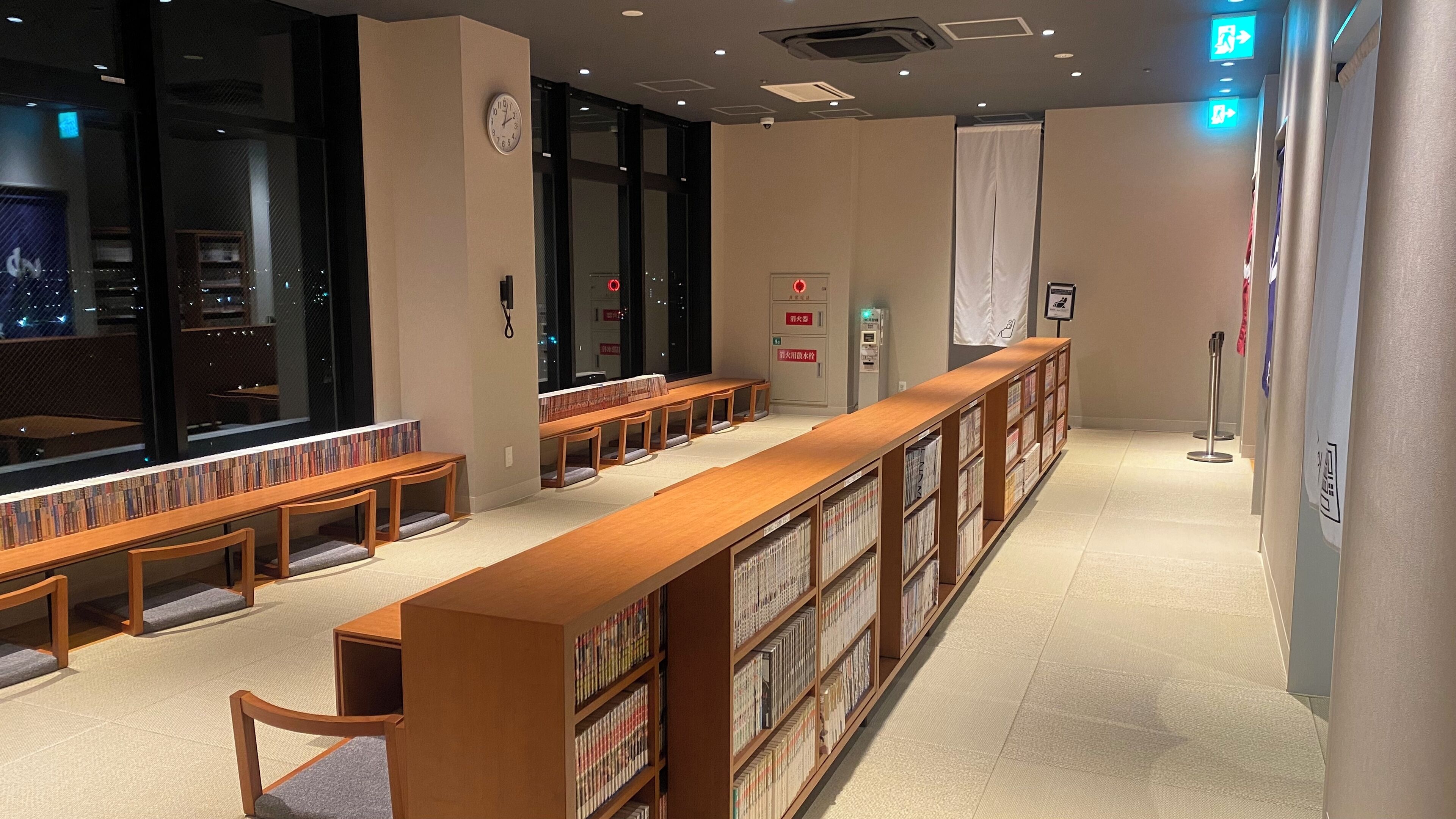 A library with wooden tables and bookshelves, large windows, and a clock on the wall.