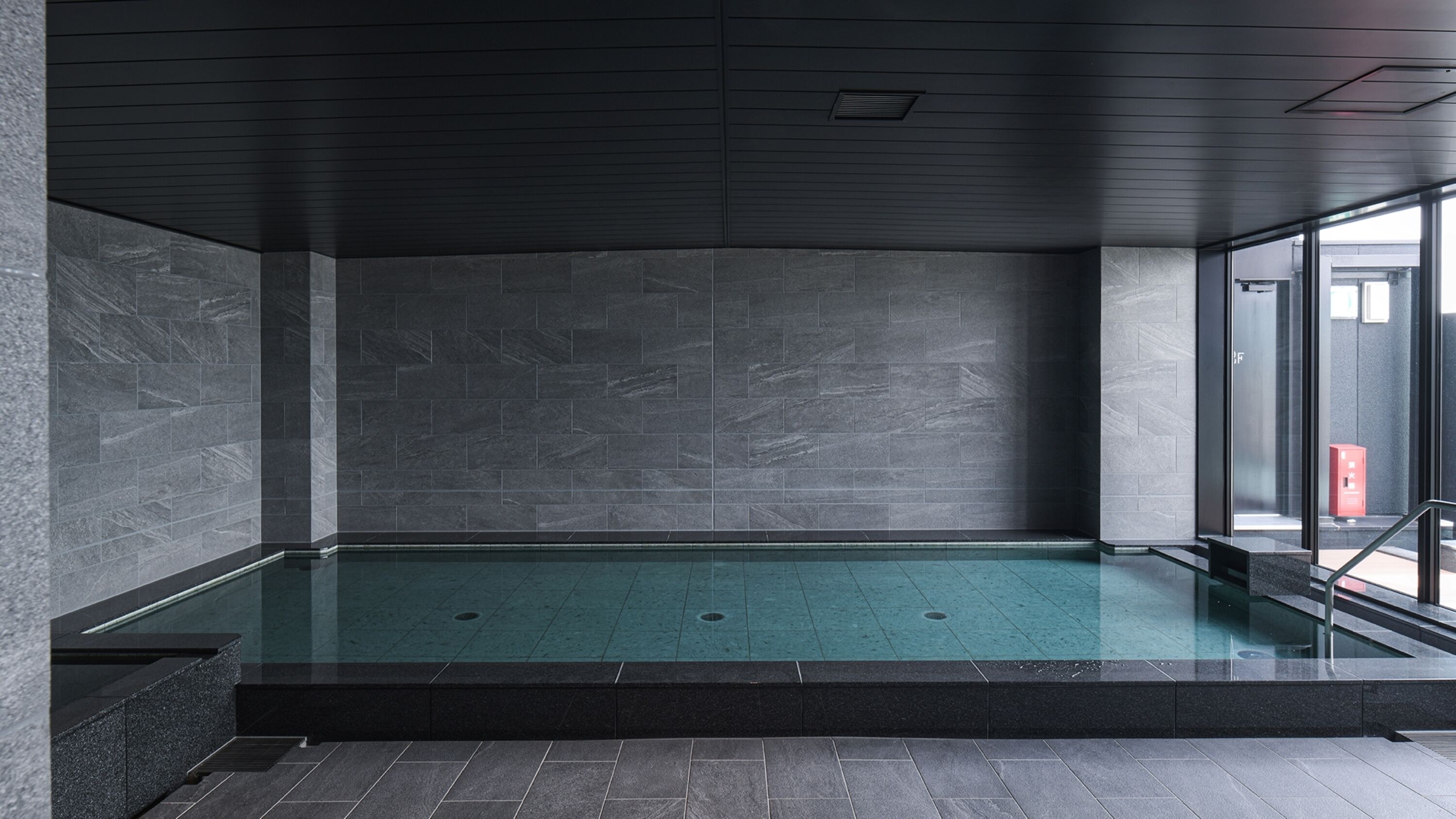 Una piscina interior moderna con paredes de azulejos oscuros y una barrera de cristal.