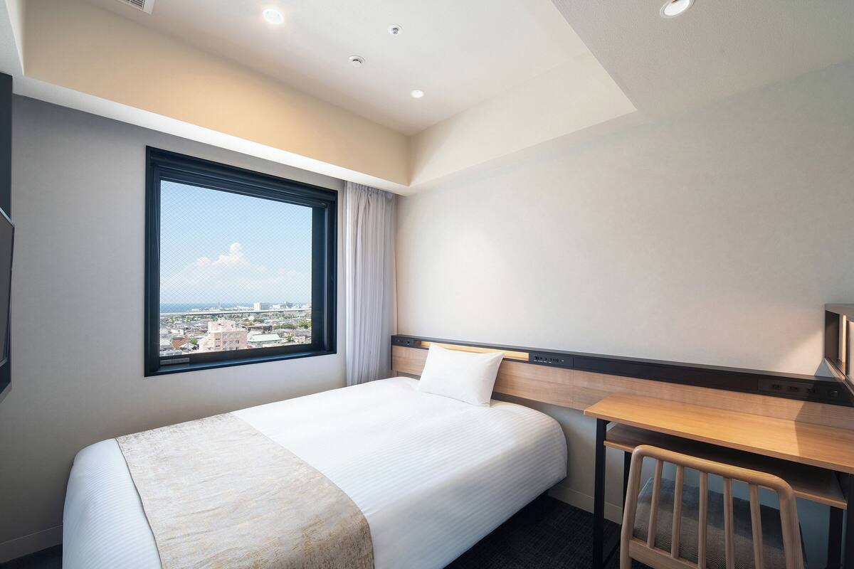 A hotel room with a bed, a desk, a chair, a window with a view, and a ceiling with recessed lighting.