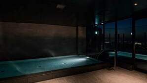 Un espace spa moderne doté d’une grande baignoire, offrant une vue sur la ville la nuit, et présentant un aménagement intérieur sombre et minimaliste.