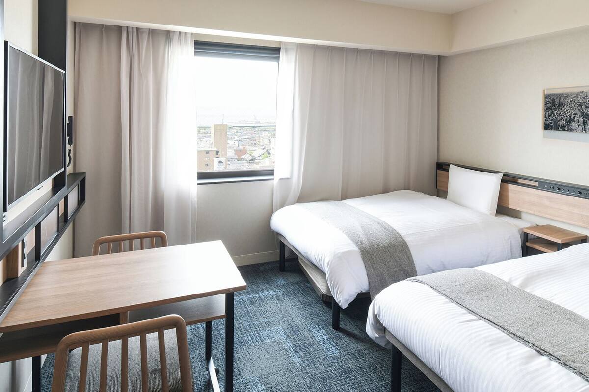 A hotel room with two beds, a desk, a chair, a TV, and a window with curtains.