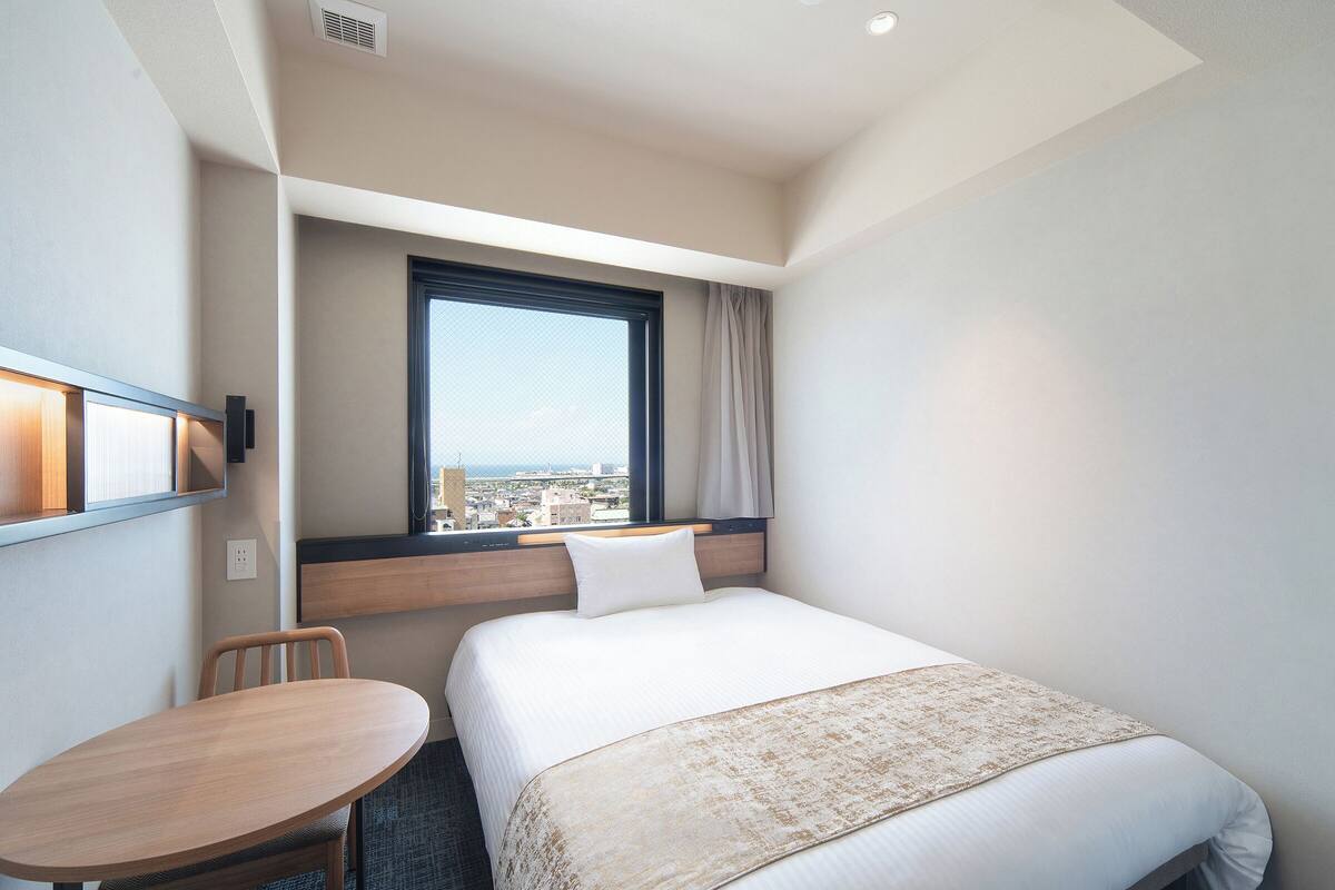 A hotel room with a bed, a desk, a chair, a window with a city view, and a wall-mounted air conditioning unit.