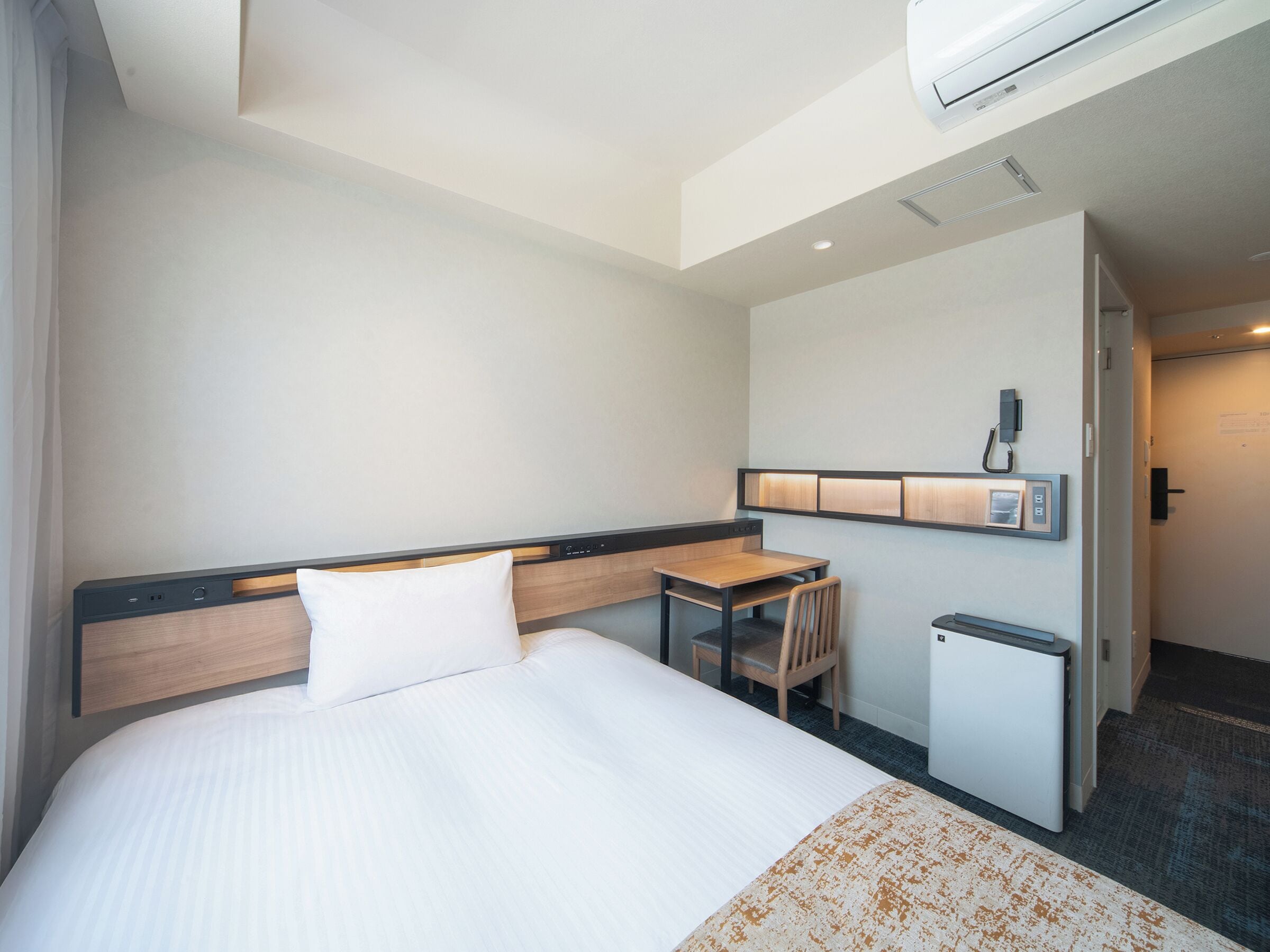 A hotel room with a bed, a desk, a chair, a small table, and a wall-mounted phone.