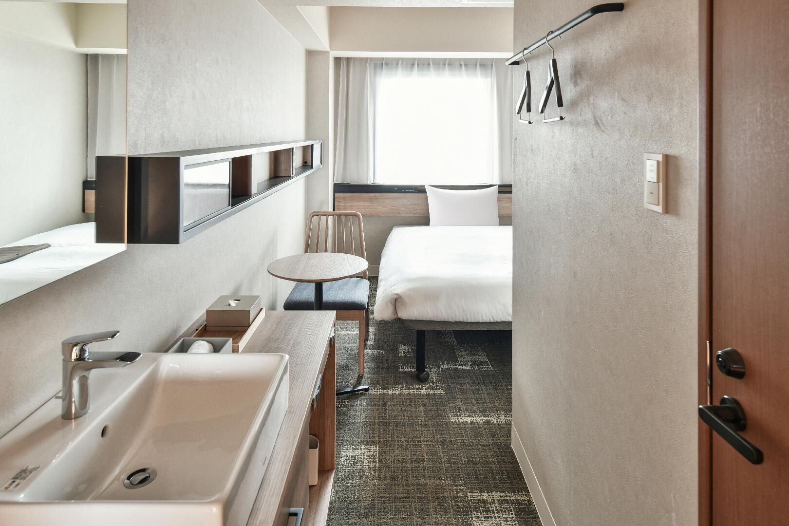A hotel room with a bed, a small round table, a chair, a sink, and a mirror.