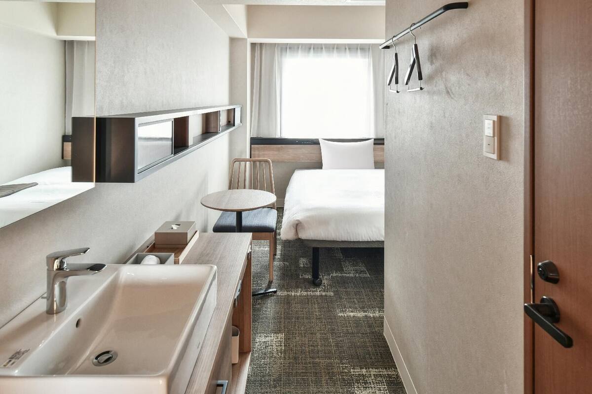 A hotel room with a bed, a small round table, a chair, a sink, and a mirror.