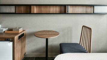 A compact hotel room with a bed, a small round table, a chair, a desk with a computer, and a built-in shelf with storage compartments.