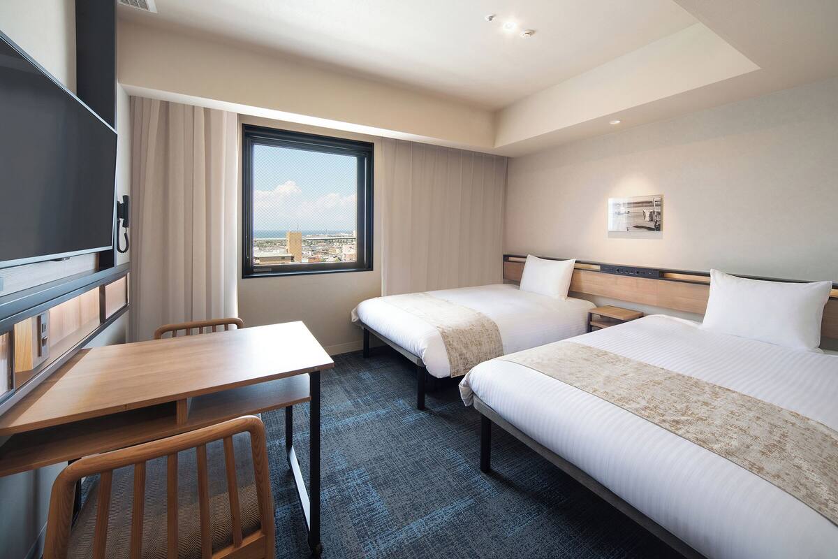 A hotel room with two beds, a desk, a chair, a TV, and a window with a city view.