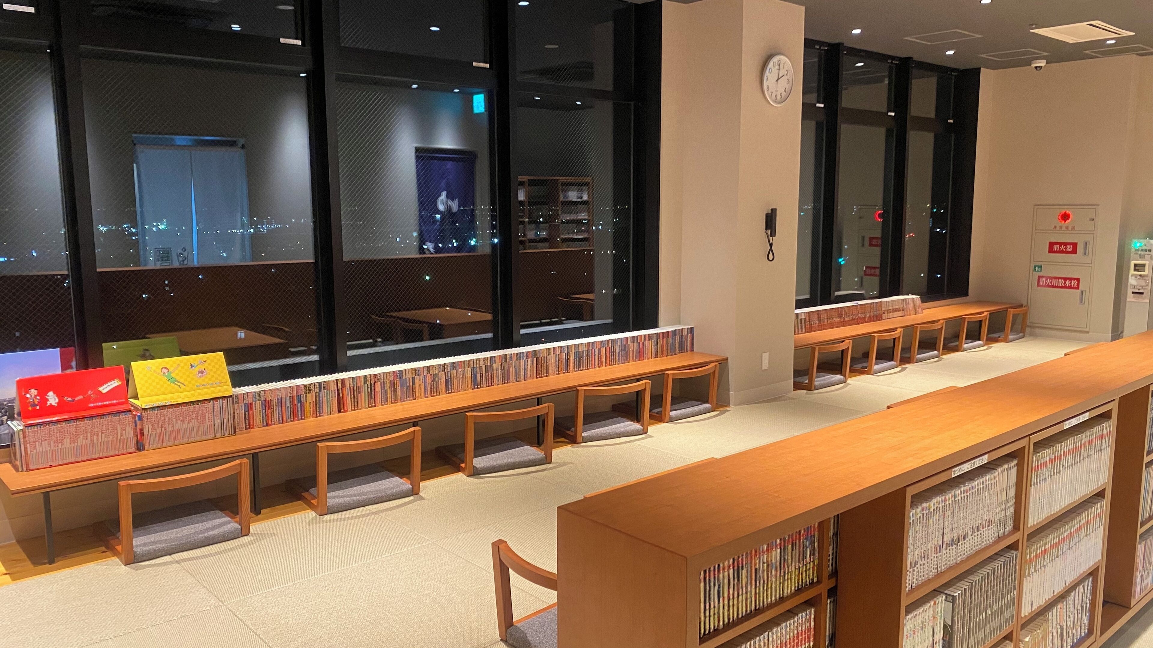 A library with bookshelves, reading tables, and a city view through large windows.