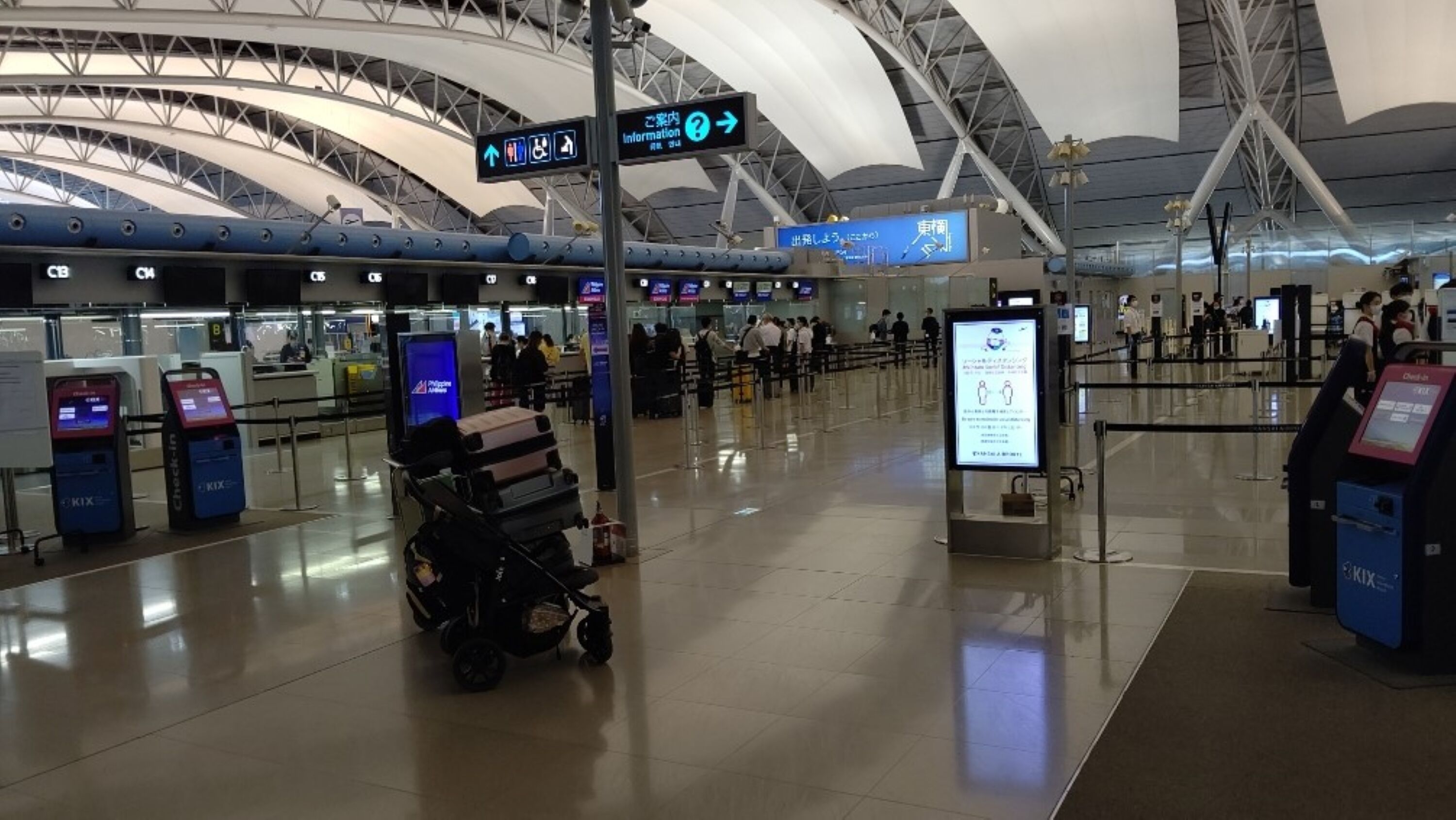 A spacious airport terminal with multiple check-in counters, a luggage trolley, and directional signage.