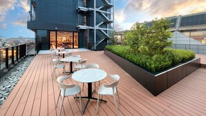 Une terrasse sur le toit avec des espaces assise extérieurs, un bâtiment moderne doté d’une façade en verre et un jardin aménagé.