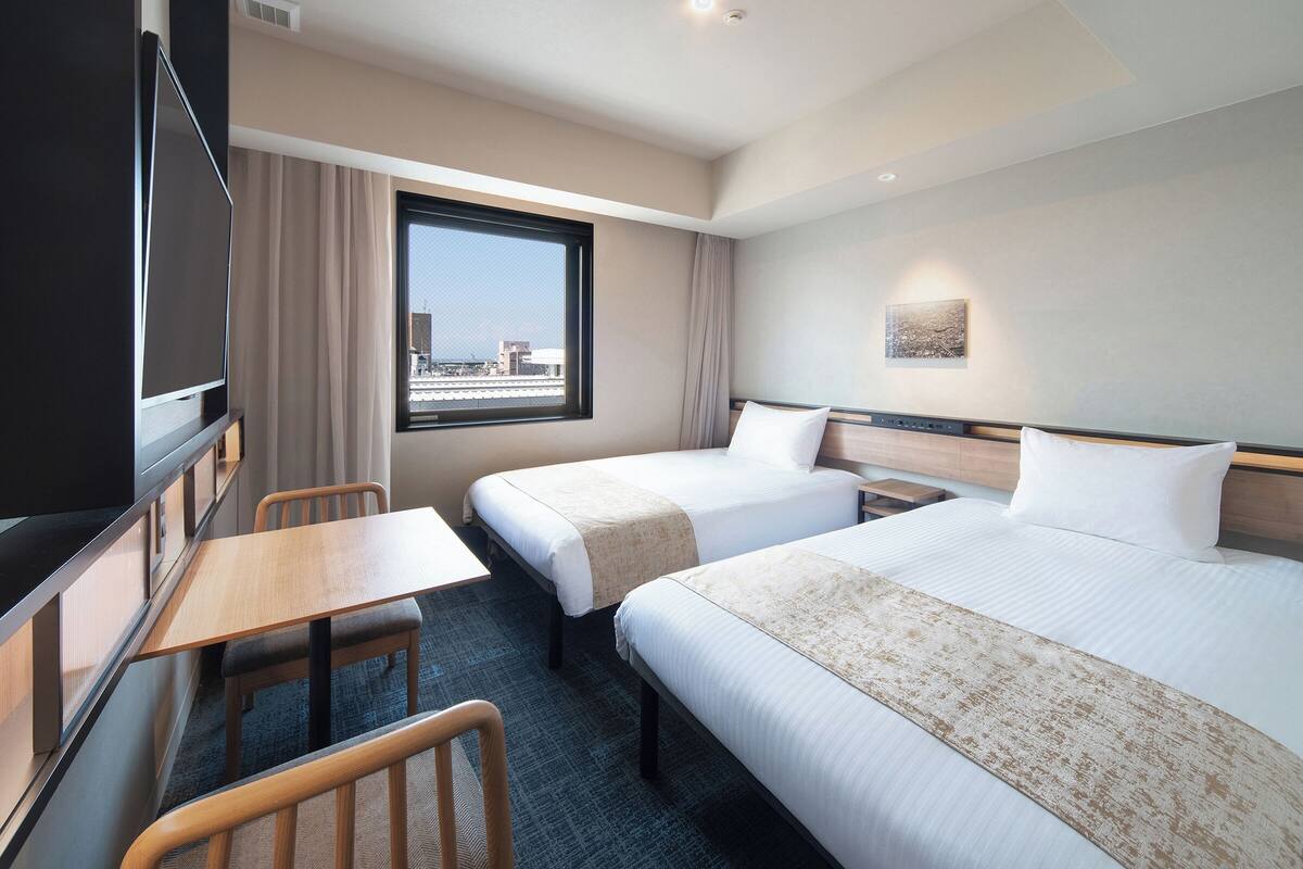 A hotel room with two beds, a desk, a chair, a TV, and a window with a city view.