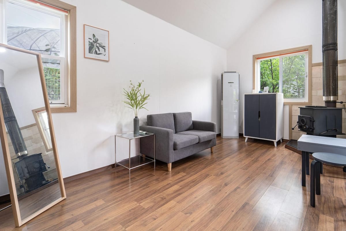 Una sala moderna con un sofá gris, una mesita auxiliar con un florero, un armario alto y una zona de cocina con estufa y fregadero.