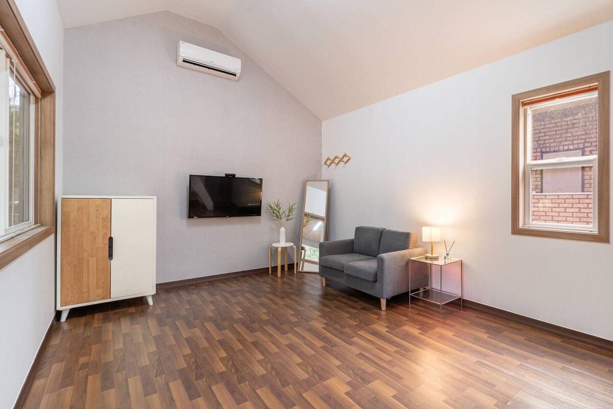 Una sala de estar moderna con piso de madera, un sofá gris, un televisor, un espejo y una pequeña mesa con una lámpara.
