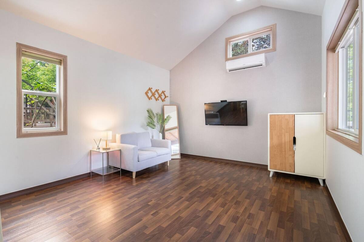 Una sala de estar moderna con piso de madera, un sofá blanco, una mesita de noche con una lámpara, un televisor montado en la pared y una ventana con vista a follaje.