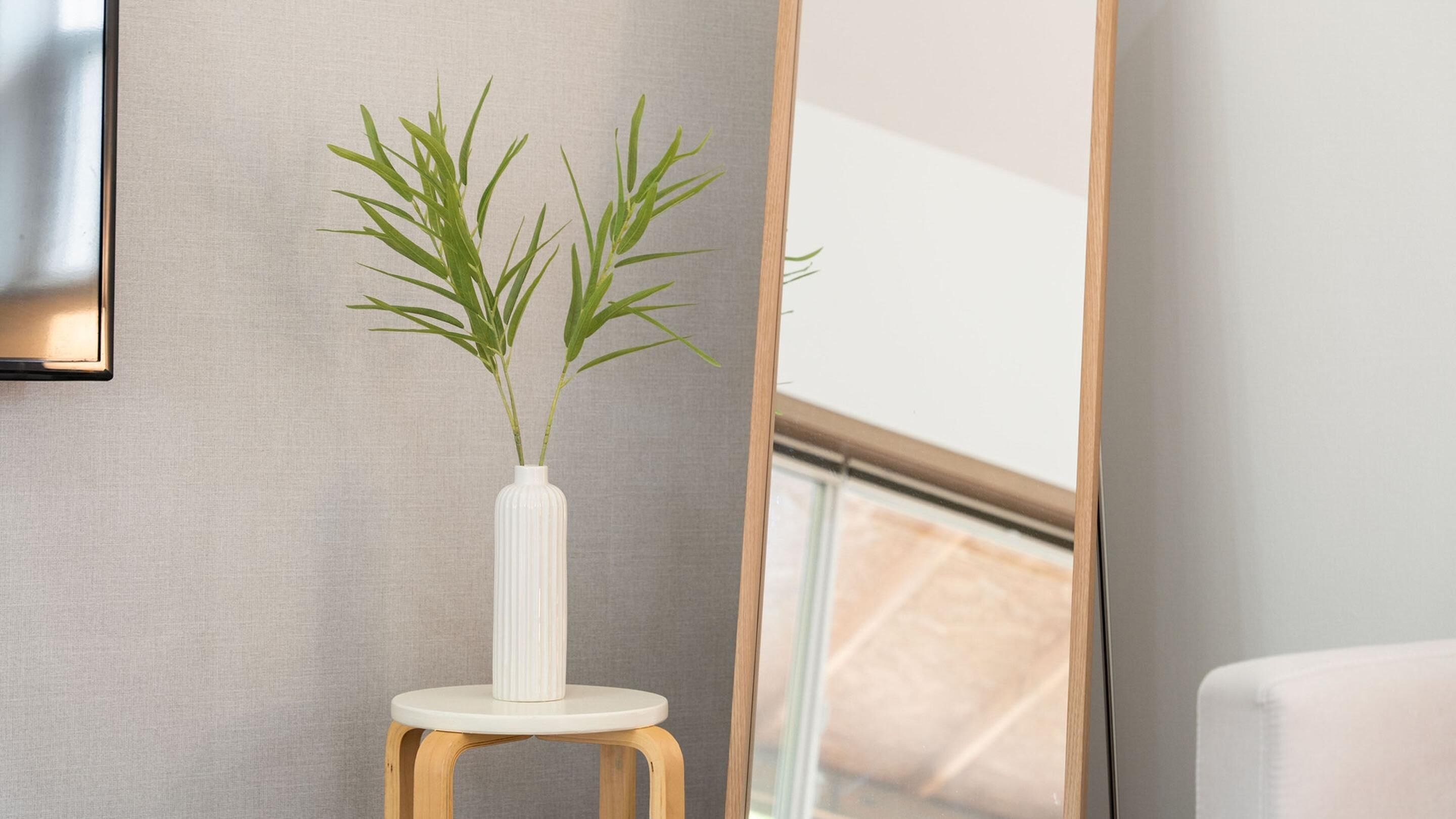 Un florero blanco con plantas verdes sobre un taburete de madera pequeño frente a una pared gris.